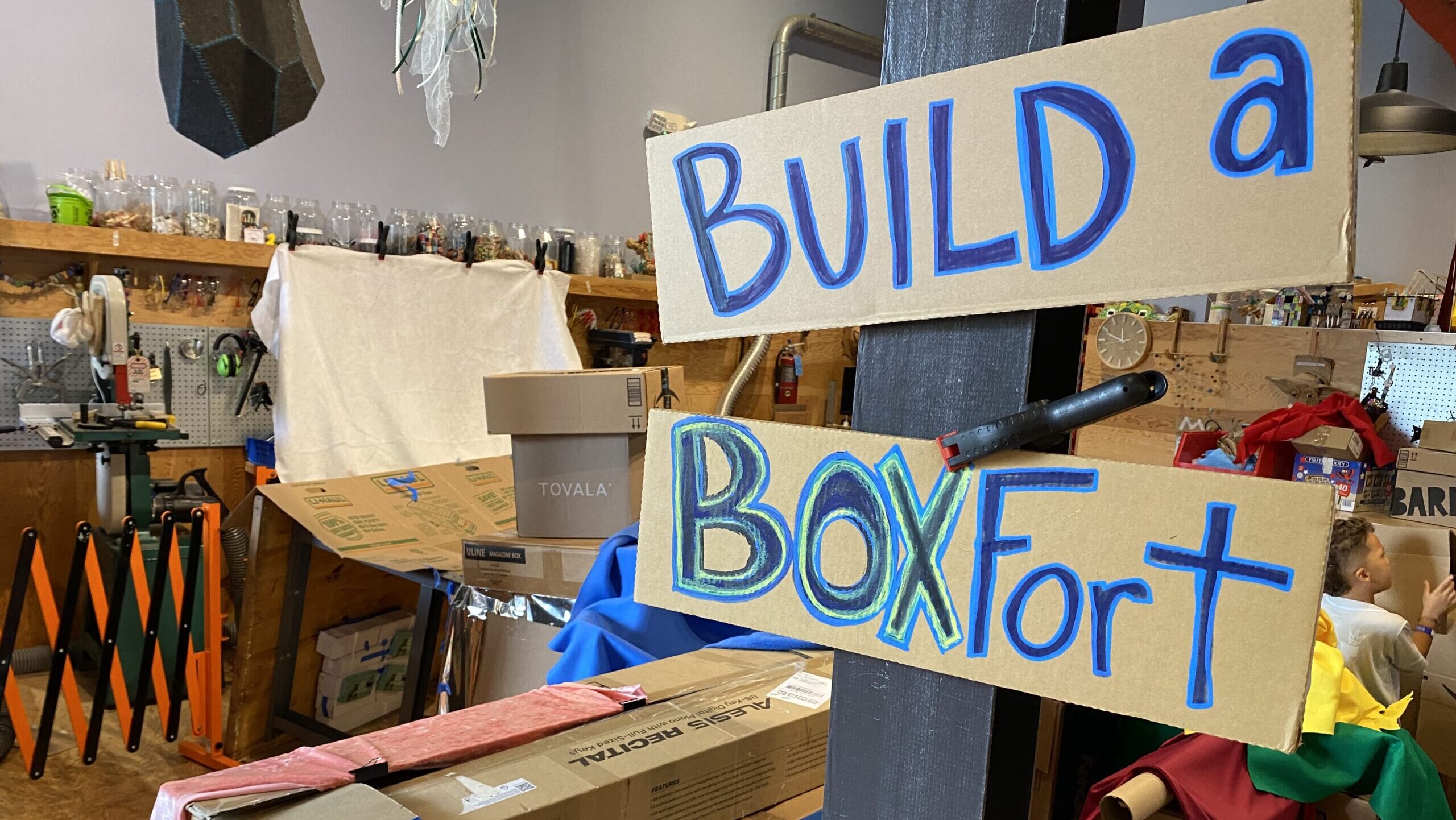 A sign reading "Build a Box Fort" in the foreground, with many types of boxes and sheets, etc, in the background