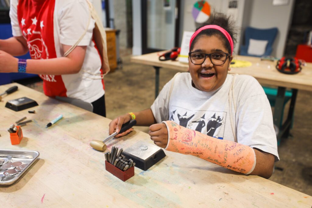 A middle school student smiles a the camera. She is using a hammer and bench block to stamp metal.