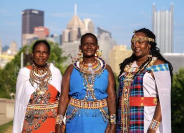 Three women wearing beaded jewelry stand in front of Pittsburgh's skyline