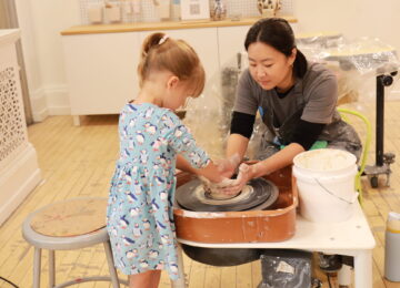 educator helps young girl mold clay on a wheel