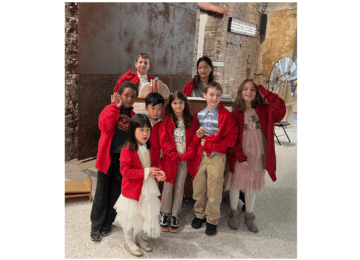 A group of children in red sweater stand around a piano