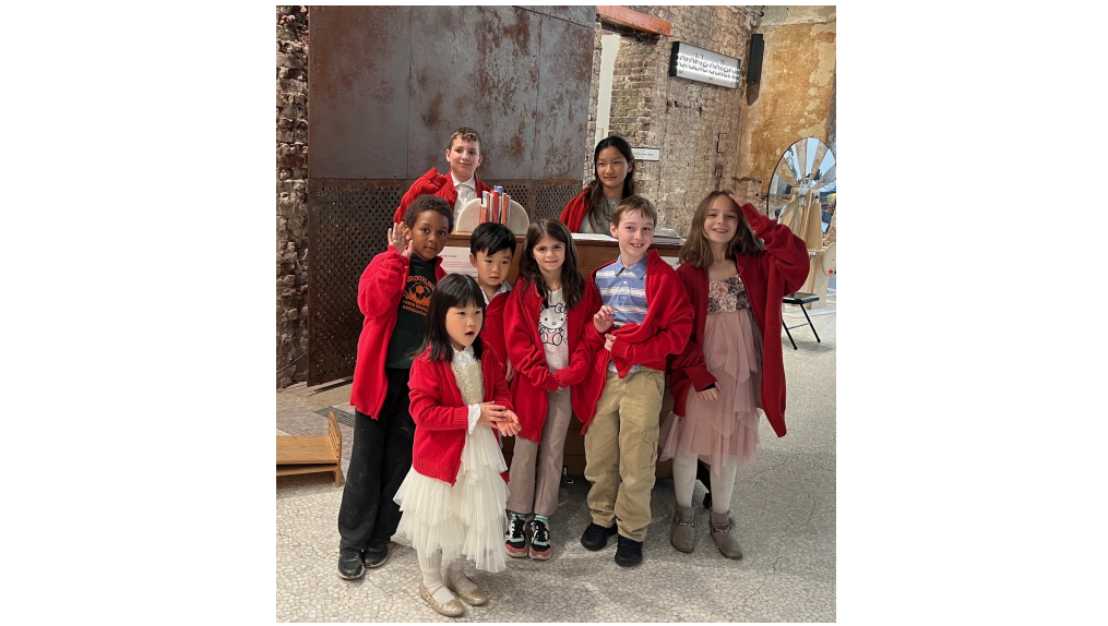 A group of children in red sweater stand around a piano