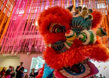 A colorful Chinese lion puppet dances in front of a large crowd in the Art Studio.
