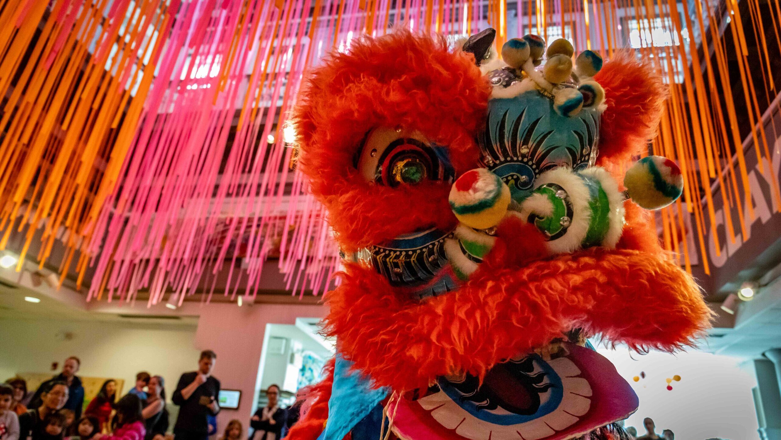 A colorful Chinese lion puppet dances in front of a large crowd in the Art Studio.