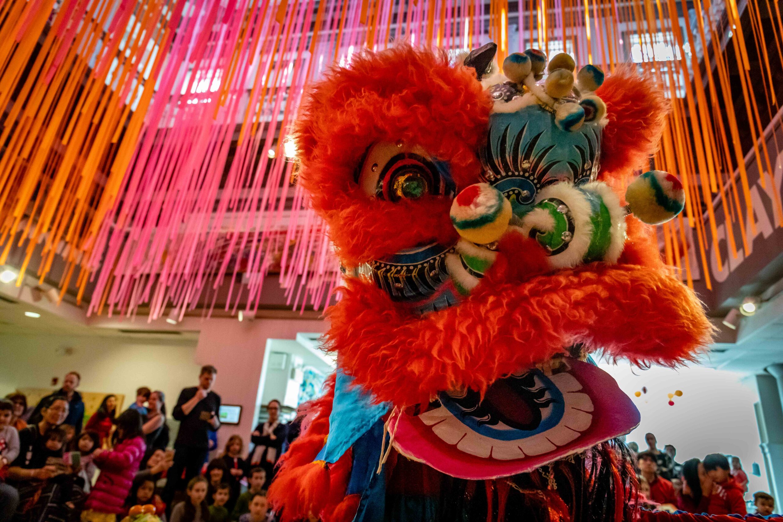 A colorful Chinese lion puppet dances in front of a large crowd in the Art Studio.