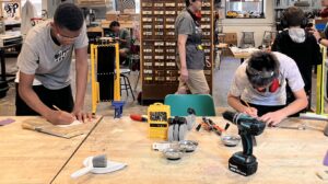 Several youth use tools to plan and mark wood at a worktable. There are tools in front of them.