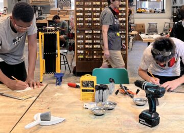Several youth use tools to plan and mark wood at a worktable. There are tools in front of them.