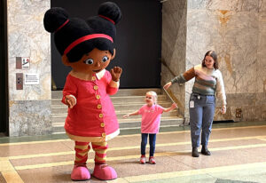 Costume of character Miss Elaina from Daniel Tiger's Neighborhood greeting a young girl with an adult woman nearby