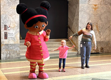 Costume of character Miss Elaina from Daniel Tiger's Neighborhood greeting a young girl with an adult woman nearby