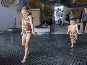 two children in bathing suits running across an indoor water play area